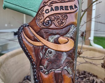 May include: A brown leather holster with intricate carvings and a bull's head design. The holster is personalised with the name "CABRERA".