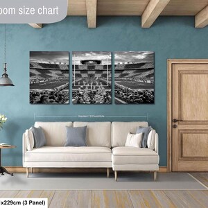 The image is a living room with a white sofa and a wooden door. Above the sofa, there is a large black and white photograph of a stadium, which appears to be a sports stadium. The photograph is divided into three panels, and the wall behind the sofa has a blue color. The living room is described as having a spacious and modern design, with a focus on the seating area and the large wall art.