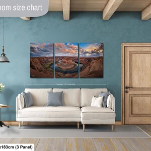 The image is a living room with a large, three-panel landscape painting hanging on the wall. The painting depicts a scenic view of a desert landscape with a river winding through it. The room has a white sofa and a wooden door, creating a cozy and inviting atmosphere.