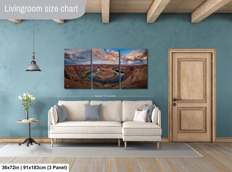 The image is a living room with a large, three-panel landscape painting hanging on the wall. The painting depicts a scenic view of a desert landscape with a river winding through it. The room has a white sofa and a wooden door, creating a cozy and inviting atmosphere.