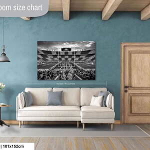 a living room with a large black and white photograph of a stadium on the wall, a white sofa, a wooden door, and a small table with a vase of flowers.
