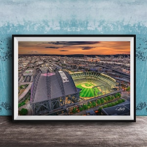 May include: Framed aerial photograph of a baseball stadium at sunset. The stadium has a retractable roof and a green field. The sky is a mix of orange and yellow hues. The stadium is surrounded by city buildings.