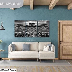 The image is a living room with a large wall-mounted painting depicting a stadium scene. The painting is divided into three panels, with the largest panel covering the entire wall. The room has a white sofa, a wooden door, and a small table with a vase of flowers. The wall is painted in a light blue color, and the floor is made of wood. The painting is described as a "Livingroom size chart" and is shown to be 36 square feet in size.