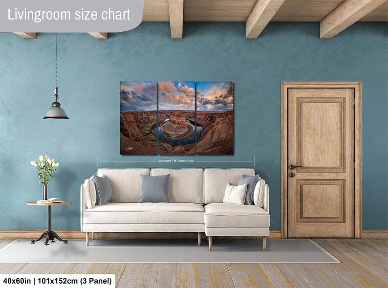 The image is a living room with a large, three-panel landscape painting hanging on the wall. The painting depicts a scenic view of a canyon with a river winding through it. The room has a white sofa and a wooden door, creating a cozy and inviting atmosphere.