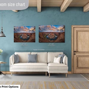 a living room with a white sofa and a wooden door. Above the sofa, there are two framed landscape photographs hanging on the wall, depicting a scenic view of a canyon or river. The wall has a blue color, and there is a lamp hanging from the ceiling. The living room appears to be well-lit and decorated, with a focus on the artwork on the wall.