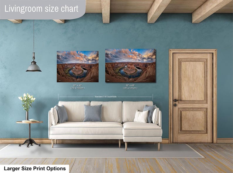a living room with a white sofa and a wooden door. Above the sofa, there are two framed landscape photographs hanging on the wall, depicting a scenic view of a canyon or river. The wall has a blue color, and there is a lamp hanging from the ceiling. The living room appears to be well-lit and decorated, with a focus on the artwork on the wall.