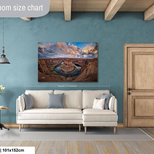 a living room with a large wall painting depicting a scenic landscape, including a river and mountains. The painting is hung above a white sofa, and there is a wooden door and a small table with a vase of flowers in the room.