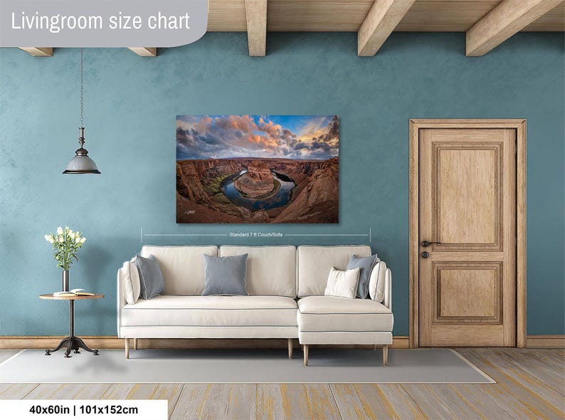 a living room with a large wall painting depicting a scenic landscape, including a river and mountains. The painting is hung above a white sofa, and there is a wooden door and a small table with a vase of flowers in the room.