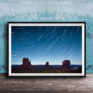 May include: Framed photograph of Monument Valley at night, with star trails against a dark blue sky. The image displays the famous sandstone buttes in silhouette, with a signature in the bottom right corner.