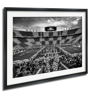 A black and white photograph of a football stadium with a large crowd of spectators, a large scoreboard, and a marching band performing on the field.