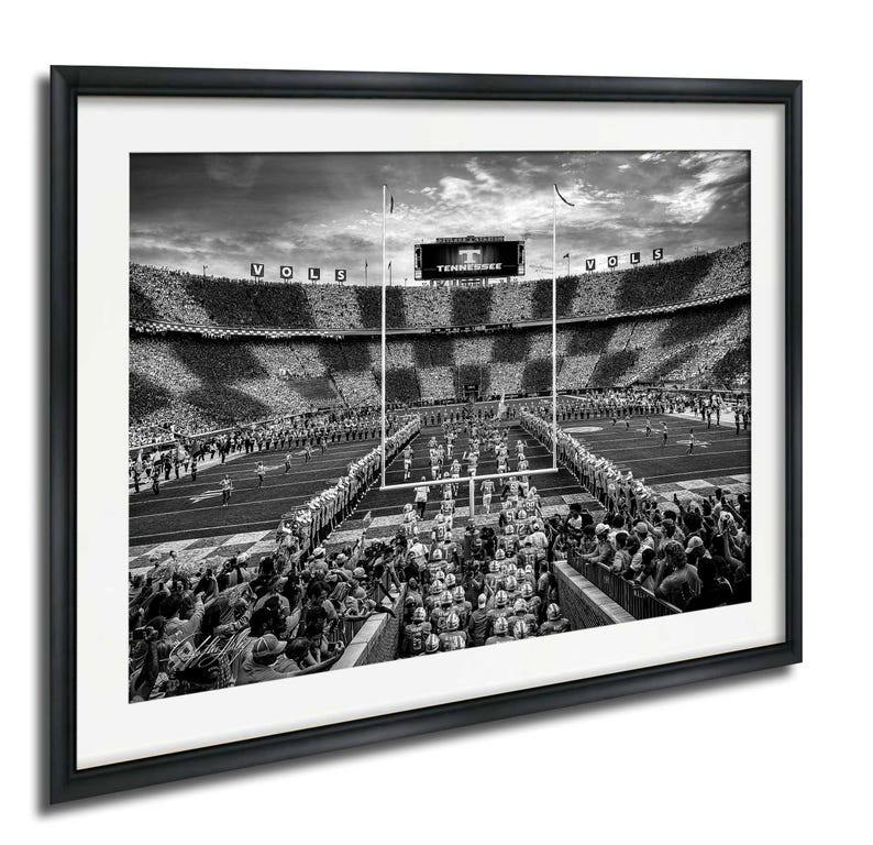 A black and white photograph of a football stadium with a large crowd of spectators, a large scoreboard, and a marching band performing on the field.