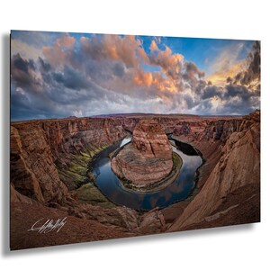 a stunning natural landscape with a winding river surrounded by towering red rock formations. The sky above is filled with clouds, creating a dramatic backdrop for the scene. The photograph is taken from a high vantage point, offering a panoramic view of the canyon and the river.