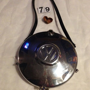 Op de afbeelding: Een ronde, zilverkleurige handtas met een zwarte riem. De tas heeft het Volkswagen-logo in het midden. Het nummer "79" wordt weergegeven op een kleine witte kaart naast een klein, hartvormig object.