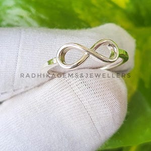 May include: A silver-colored ring in the shape of an infinity symbol. The ring is displayed on a white surface with green foliage in the background. The text "RADHIKAGEMS&JEWELLERS" is visible on the surface.