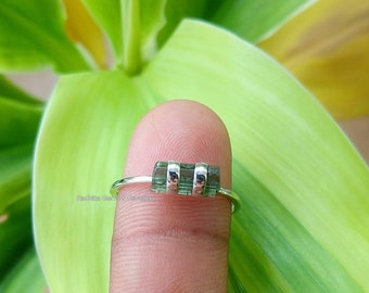 May include: A silver ring with three green gemstones set in a row. The ring is on a finger.