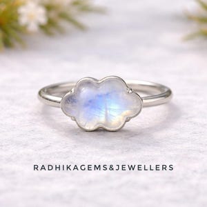 Puede incluir: Un anillo de plata con una piedra preciosa en forma de nube con tonos iridiscentes azules y blancos. El anillo tiene una banda delgada y pulida. El texto "RADHIKAGEMS&JEWELLERS" está impreso debajo.