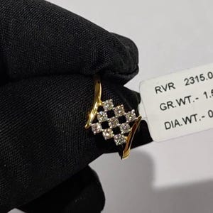 May include: A gold ring with a diamond-studded, checkered design. The ring features a series of small, clear diamonds set in a square pattern. The ring is held against a black background, with a price tag visible.