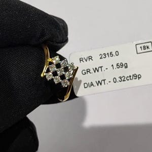May include: A gold ring with a diamond-studded, checkered design. The ring features a cluster of small, round diamonds set in a geometric pattern. The ring is held against a dark background, with a tag that reads "18k".