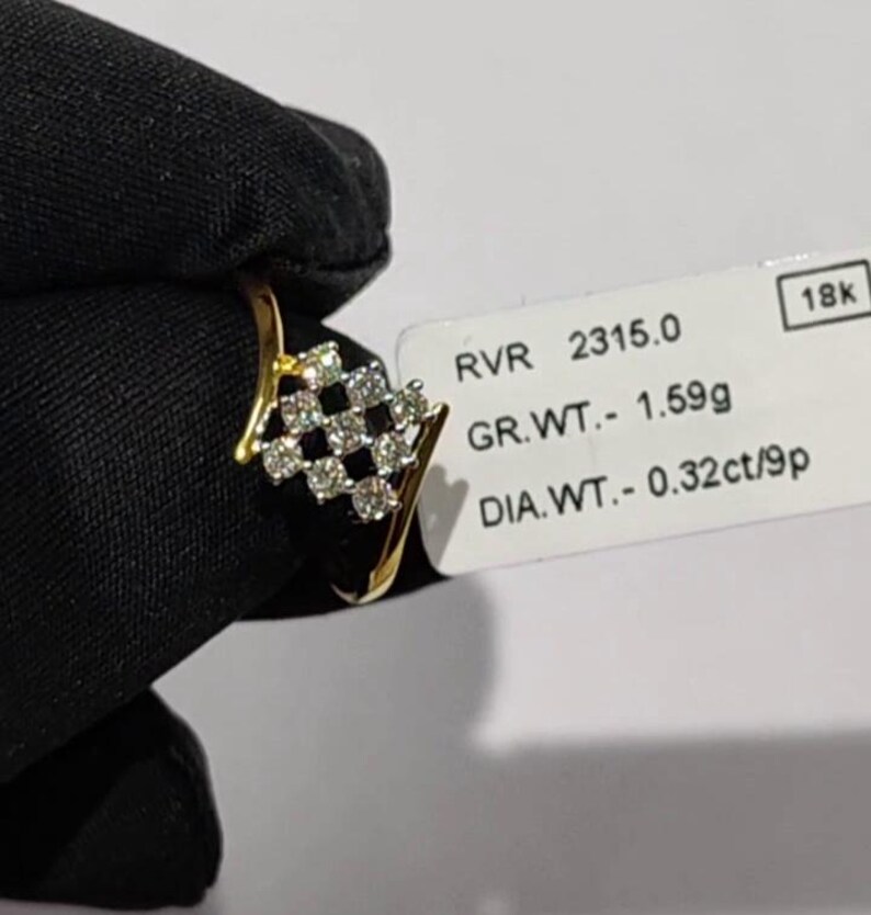 May include: A gold ring with a diamond-studded, checkered design. The ring features a cluster of small, round diamonds set in a geometric pattern. The ring is held against a dark background, with a tag that reads "18k".
