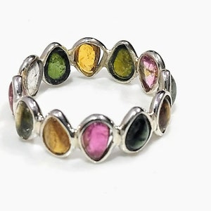 Puede incluir: Un anillo de plata con una banda de piedras preciosas de colores. Las piedras están dispuestas en una fila alrededor de la banda y todas tienen diferentes formas y tamaños. Las piedras son una mezcla de rosa, verde, amarillo y negro.