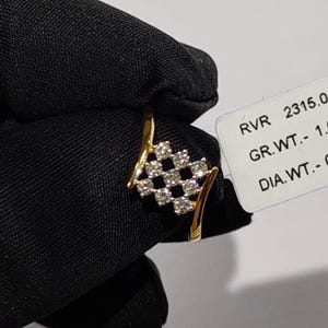 May include: A gold ring with a diamond-studded checkerboard design. The ring has a curved band and a cluster of diamonds set in a square pattern. The ring is held against a black background.