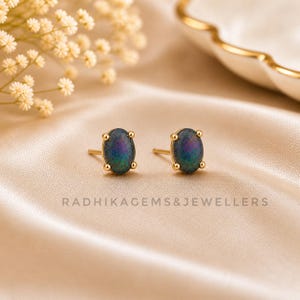 Könnte beinhalten: Goldene Ohrstecker mit ovalen, irisierenden schwarzen Edelsteinen. Die Ohrringe sind in Gold gefasst und werden auf einer satinartigen Oberfläche präsentiert. Der Text "RADHIKAGEMS&JEWELLERS" ist sichtbar.