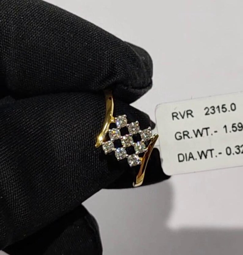 May include: A gold ring with a diamond-studded square design. The ring features a series of small, clear diamonds set in a grid pattern. The ring is held against a black background, with a tag visible that includes the text "RVR 2315.0".