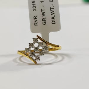 May include: A gold ring with a cluster of small, clear stones in a square pattern. The ring has a split shank design and is set against a white background. A tag with text is visible.