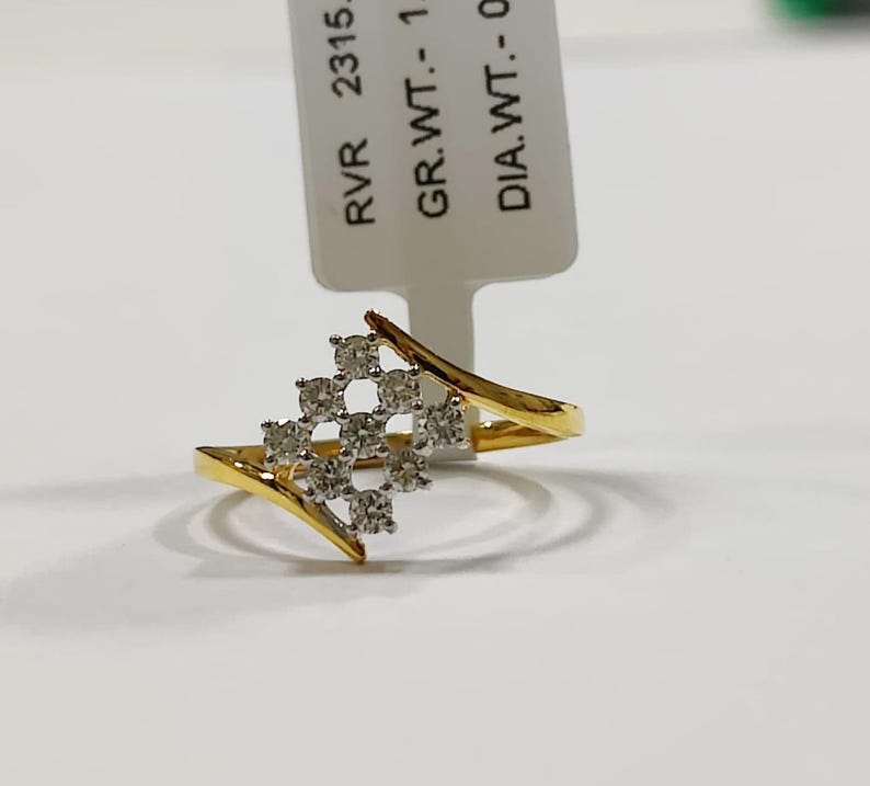 May include: A gold ring with a cluster of small, clear stones in a square pattern. The ring has a split shank design and is set against a white background. A tag with text is visible.