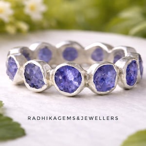 May include: A silver ring featuring a series of oval, rough-cut, blue-purple gemstones. The gemstones are set in individual silver bezels, creating a continuous band. The ring is displayed against a soft, white background with the text "RADHIKAGEMS&JEWELLERS".