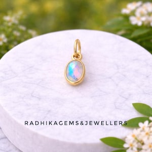 Può includere: Un pendente in oro con una pietra preziosa di opale ovale. L'opale mostra colori iridescenti di blu, rosa e verde. Il pendente è incastonato in una lunetta d'oro e ha un piccolo anello per una catena. Il testo "RADHIKAGEMS&JEWELLERS" è in basso.