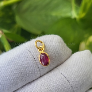 May include: A gold pendant with a vibrant red oval-shaped gemstone. The pendant is suspended from a small loop and is set against a soft, neutral background. The gemstone is the focal point, showcasing its rich color and polished surface.