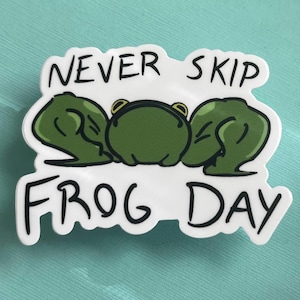 May include: White sticker with a green frog illustration and the text "Never skip frog day".