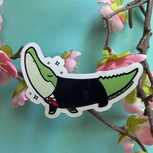 May include: A green alligator wearing a black suit and a red tie. The alligator is smiling and has a sparkle effect around it. The sticker says "Ceno the Crocodile".