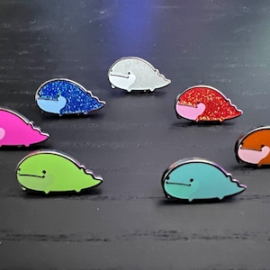 May include: Eight enamel pins shaped like whales in various colors, including pink, blue, green, red, and white. The pins have glitter accents and are arranged on a dark surface.