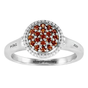 May include: A silver ring with a round center stone set in a halo of smaller stones. The center stone is a cluster of brown diamonds. The ring is engraved with "MRA" on one side and "2021" on the other. The ring is also engraved with "silvergiftjewellery".