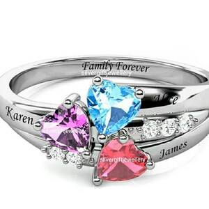Op de afbeelding: Zilveren ring met drie hartvormige edelstenen in blauw, roze en rood. De ring is gegraveerd met de woorden "Family Forever" en de namen "Karen", "Alice" en "James".