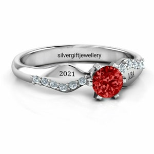 May include: A silver ring with a red gemstone and a row of smaller clear gemstones. The ring is engraved with "silvergiftjewellery", "2021", and "MBA".