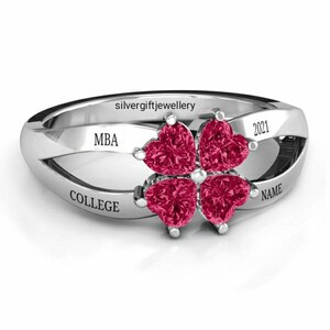 May include: Silver ring with four red heart-shaped stones set in a clover design. The ring is engraved with the words "MBA", "COLLEGE", "NAME", and "2021".