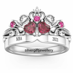 May include: A silver ring with a crown design featuring two red heart-shaped stones and smaller red stones on the sides. The band is engraved with "MBA" on one side and "2021" on the other. The ring is made by silvergiftjewellery.