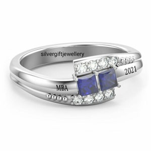 May include: Silver ring with two blue square gemstones and a row of smaller clear gemstones. The ring is engraved with "MBA" and "2021".