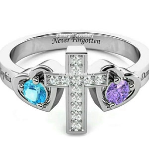 May include: A silver ring with a cross design, two heart-shaped settings with blue and purple gemstones, and the inscription "Never Forgotten". The ring is engraved with the names "Sophia" and "Daniel".
