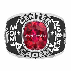 Puede incluir: Un anillo de clase de plata con fondo negro. El anillo presenta las palabras "Center Academy Ka-Ron" y el año "2021". Una piedra preciosa roja está engastada en el centro del anillo.