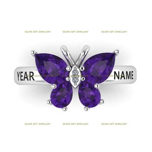 May include: A silver ring with a purple butterfly design. The butterfly has four wings with pear-shaped purple gemstones. The ring band is engraved with the words "YEAR" and "NAME".