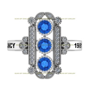 May include: A silver ring with three blue gemstones set in a decorative frame. The ring is inscribed with the year "1981" and the word "ANCY".