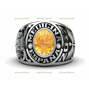 Puede incluir: Un anillo de plata con una banda de esmalte negro y una piedra preciosa redonda grande de color amarillo. El anillo tiene las palabras "MEDICINA ESPAÑA" en letras negras.