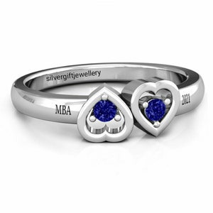 May include: A silver ring with two heart-shaped settings, each containing a blue gemstone. The ring is engraved with "MBA" and "2021".