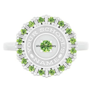 May include: A silver ring with a circular design. The ring features a central green gemstone and is encircled by smaller green gemstones. The words "YOUR SCHOOL'S NAME" are inscribed around the center. The ring is a bright, shiny silver color.