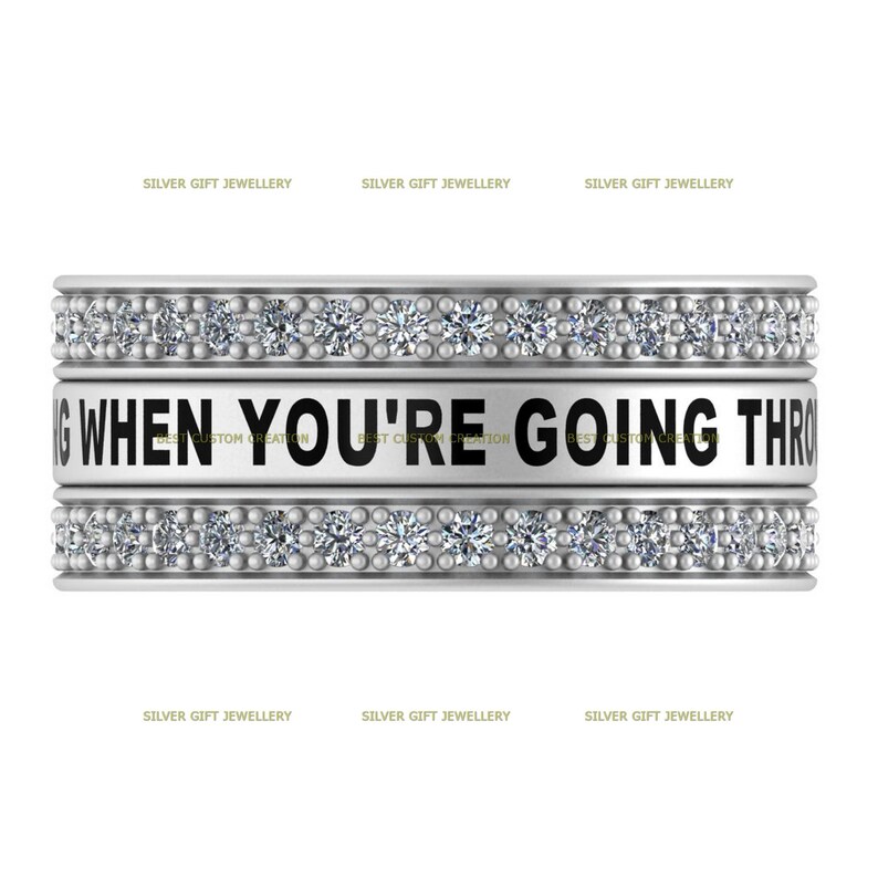 Op de afbeelding: Een zilveren ring met een zwarte band waarop "WHEN YOU'RE GOING THROUGH" staat en die is versierd met kleine, heldere stenen.