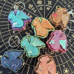 Può includere: Una collezione di portachiavi a forma di drago colorati. Ogni portachiavi presenta un drago con ali dettagliate e una finitura metallica. I portachiavi sono in vari colori, tra cui verde acqua, arancione e viola, e sono attaccati ad anelli argentati.